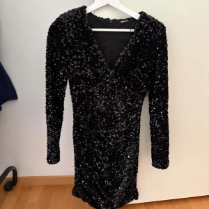 Svart paljettklänning  - Supersnygg svart kort paljettklänning från Gina tricot med v-ringning och långa ärmar. Tight passform men ändå stretchig, axelvaddar finns också. I strl S, endast använd en gång.