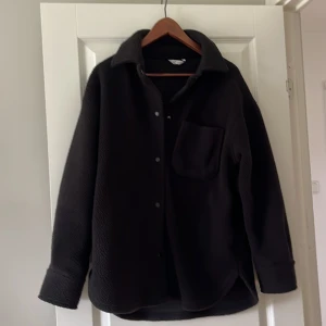 Svart oversized overshirt från C - Svart oversized overshirt från C i storlek XS. Skjortan har en mjuk, fleece-liknande teddy känsla, klassisk krage, knappstängning framtill och en bröstficka. Perfekt för lager-på-lager och chill stil.