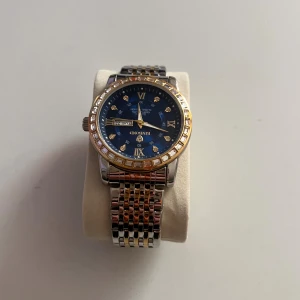 Lyxig klocka - Stilren klocka med blå urtavla, guldfärgade romerska siffror och stenar som index. Boetten är silverfärgad med guldfärgad ring runt urtavlan. Länkarmbandet är i tvåtonad silver och guld, vilket ger en lyxig känsla. Klockan har dag- och datumvisning.