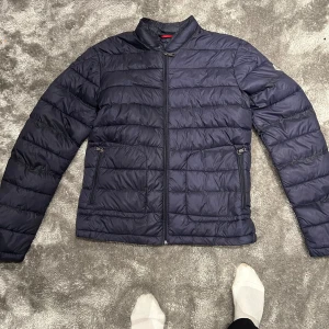 Mörkblå dunjacka från Moncler - Snygg mörkblå dunjacka från Moncler med klassisk quiltad design och diskret logga på ärmen. Jackan har dragkedja framtill och två fickor med dragkedja. Perfekt för kyliga dagar och ger en clean look.