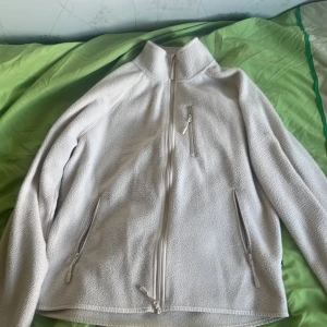 Vit fleecejacka med dragkedjor  -  Mysig vit fleecejacka med hög krage och tre dragkedjeförsedda fickor framtill. Jackan har en enkel och stilren design, perfekt för lager-på-lager och chill dagar. Långärmad och med mjukt material som håller dig varm.  