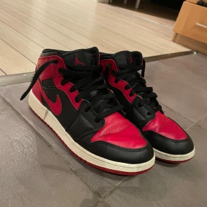 Nike Air Jordan 1 Mid Bred EUR 40 - Säljer ett par Nike Air Jordan 1 Mid i den klassiska Bred-färgen. Skorna har svart och rött skinn, svart swoosh och röda detaljer. Skorna har tyvärr ett litet hål innuti skon.Ikonisk siluett med snörning och vit mellansula. Perfekta för dig som gillar streetwear och sneakers med attityd.