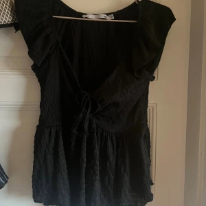 Svart omlottopp med volang från 157 - Supersöt svart omlottopp från 157 i storlek XS. Toppen har volangdetaljer vid axlarna, knytning framtill och är tillverkad i ett lätt, krinklat tyg. Perfekt för dig som gillar feminina och trendiga plagg med snygg passform och extra detaljer.