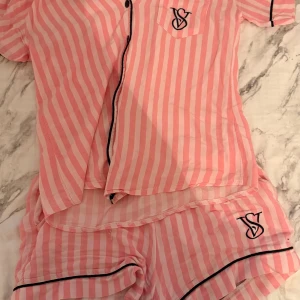 Randiga pyjamasshorts från Victoria's Secret - Säljer ett par supergulliga pyjamasshorts från Victoria's Secret i rosa och vitt med ränder. Shortsen har svarta detaljer och VS-logga broderad framtill. Perfekta för mysiga kvällar hemma. Materialet känns lätt och luftigt.