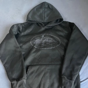 Corteiz hoodie - Svart hoodie med huva och magficka. På bröstet finns ett tryck i silver av ett fartyg inuti en oval ram. Tröjan har långa ärmar med ribbade muddar och är gjord i ett mjukt material som känns skönt mot huden.