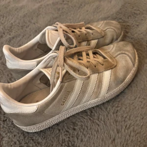 Beige Adidas Gazelle sneakers mocka - Säljer ett par klassiska Adidas Gazelle sneakers i beige mocka med vita detaljer och snörning. Skorna är lite slitna där fram men inte något som märks tydligt💕