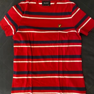 Röd randig t-shirt från Lyle & Scott - Snygg röd t-shirt från Lyle & Scott med breda marinblå och vita ränder. Klassisk rund hals och korta ärmar. På bröstet sitter den ikoniska gula fågeln broderad. Mjuk bomullskvalitet som känns skön mot huden. Perfekt för dig som gillar sportig och stilren vibe. Storlek XXL men passar som XL