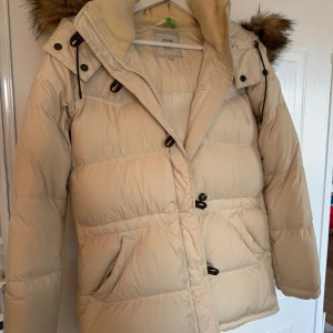 Beige dunjacka med fuskpäls från Boomerang - Snygg beige dunjacka från Boomerang med stor huva och fuskpälsdetalj. Jackan har dragkedja, knappar och två stora fickor framtill. Huvan är avtagningbar. Klassisk och stilren look som håller dig varm.