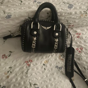  Zadig & Voltaire sunny bag xsmall - Svart handväska i skinn från Zadig & Voltaire med silvriga nitar, dubbla handtag och bred axelrem med logga. Väskan har dekorativa dragkedjor framtill och ett metallmärke med vingar. Fint skick, dustbag finns med, dock från annan Zadig väska. 🥰