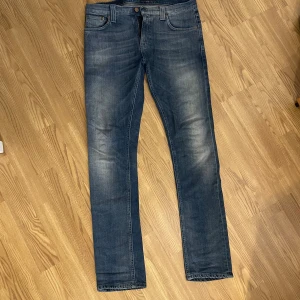 Blågrå slim fit jeans från Nudie Jeans - Säljer ett par blågrå slim fit jeans från Nudie med klassisk femficksdesign och snygga slitningar på låren. Jeansen har normal midja och raka ben med en smal passform. Tillverkade i mjukt denimtyg som sitter skönt hela dagen. Modell Tube Tom, storlek 31W/32L (Modell 176cm, 70kg)