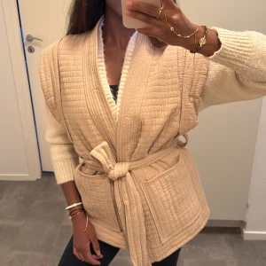 Beige quiltad väst med knytband - Superfin beige quiltad väst med breda axlar och knytband i midjan. Västen har stora fickor framtill och en mjuk, vadderad känsla 🍂🧚🏾‍♀️