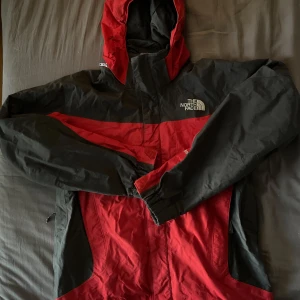 Röd och svart vindjacka från The North Face - Säljer en snygg röd och svart vindjacka från The North Face med huva och dragkedja. Jackan har sportig look, meshfoder och logga på bröstet och ryggen. Perfekt för dig som gillar streetwear och outdoor vibes.
