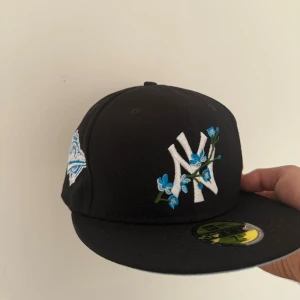 New Era keps - Säljer en svart New Era 59FIFTY Yankees keps med broderade blå blommor och vit NY-logga framtill. På sidan finns ett blått World Series 1996-märke och New Era-logga. Undersidan av skärmen är ljusblå. Perfekt för dig som vill sticka ut med snygga detaljer.