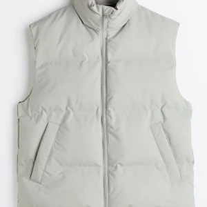 Beige dunväst från H&M, storlek L - Snygg beige dunväst från H&M i storlek L. Västen har hög krage, dragkedja framtill och två snedställda fickor. Materialet är både vattenavvisande och vindtät. Perfekt för lager-på-lager under kyliga dagar.
