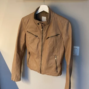 Beige skinnjacka från Cappuccini - Snygg beige skinnjacka från Cappuccini med dragkedjor framtill och vid fickor. Jackan har hög krage med knapp och en figurnära passform. 