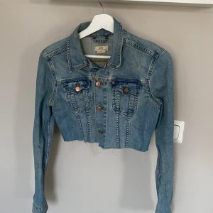 Croppad jeansjacka från &NOW, strl 36 - Snygg croppad jeansjacka från &NOW i ljusblå denim. Jackan har rå kant nertill, klassisk krage, bröstfickor med knappar och långa ärmar. 