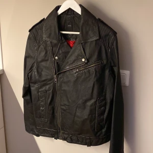 Svart skinnjacka från Emilio, stl S - Säljer en svart skinnjacka från Emilio i storlek S. Jackan har klassisk bikerstil med sned dragkedja, axelklaffar och coola detaljer som fickor med dragkedja. Röd insida som ger extra edge. 