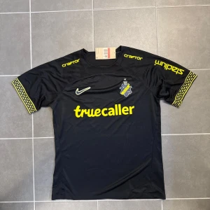 AIK svart matchtröja Nike L - AIK:s officiella svarta fotbollströja från Nike i storlek L. Tröjan har gula detaljer, klubbmärke på bröstet, sponsorloggor och unika mönstrade muddar på ärmarna. Tillverkad i lätt och ventilerande funktionsmaterial, perfekt för match eller träning.