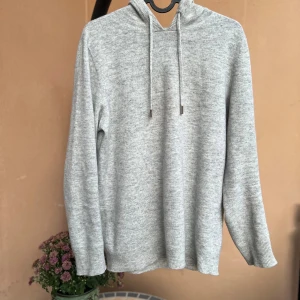 Ljusgrå stickad hoodie med huva - Mysig ljusgrå stickad hoodie med huva och dragsnören. Tröjan har lång ärm, ribbade muddar och en enkel, clean look som passar perfekt till chill dagar. Materialet är mjukt och ger en avslappnad vibe.