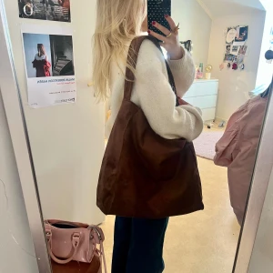 Stor brun axelväska i mocka - Säljer en stor och mjuk axelväska i brun mocka. Väskan har enkel design med bred axelrem och rymligt huvudfack, perfekt för att få plats med allt du behöver. Snygg och stilren look som funkar till många outfits.