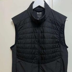 Svart quiltad dunväst från Hugo Boss - Stilren svart dunväst från Hugo Boss med quiltad framsida och baksida, hög krage och dragkedja. Västen har diskret logga på bröstet och ryggen, samt är tillverkad i lätt och vattenavvisande material. Perfekt för lager-på-lager och streetstyle.