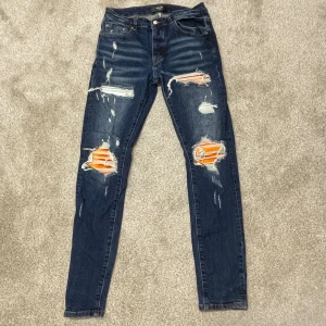 Amiri Jeans - Mörkblå skinny jeans från Amiri med kraftiga slitningar och trasiga detaljer framtill, där orange och vita ränder syns under hålen. Klassisk femficksmodell med läderpatch bak i midjan och metallogo vid bakfickan. Snygg streetstil med smal passform. Skicket på jeansen är bra och storleken är M (Pris kan diskuteras).