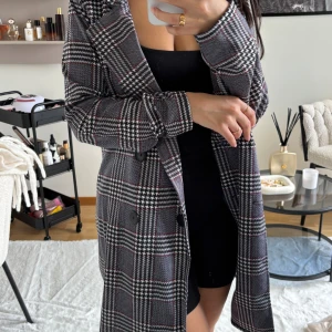 Rutig kappa i grått och svart - Snygg lång kappa med rutig design i grått, svart och röda detaljer. Klassisk krage, knappar framtill och rak passform. Perfekt för dig som vill ha en stilren och trendig look. Materialet känns som en blandning av ull och polyester.