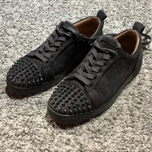 Gråa sneakers från Louboutin med nitar - Säljer ett par grå sneakers från Christian Louboutin i mocka med ikoniska röda sulor. Skorna har rund tå, snörning och coola svarta nitar på tån för en edgy vibe. Perfekta för dig som vill sticka ut med lyxig streetstyle.