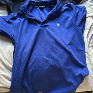 Två pikétröjor från Ralph Lauren - Säljer två klassiska pikétröjor från Ralph Lauren. En är marinblå med röd broderad logga och vita knappar, den andra är blå med vit broderad logga. Båda har kort ärm, krage och är gjorda i mjuk bomull. Perfekta för en stilren och avslappnad look. Strlk M