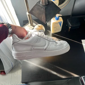 Nike Air Force 1 vita sneakers - Klassiska Nike Air Force 1 sneakers i helvitt skinn med rund tå, snörning och platt sula. Ikonisk design med perforerade detaljer på tån och broderad Nike-logga på hälen. Perfekta för dig som vill ha en clean och tidlös look.