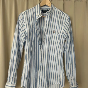 Randig skjorta från Polo Ralph Lauren - Snygg slim fit skjorta från Polo Ralph Lauren med vertikala blå och vita ränder. Perfekt för dig som gillar stilrena och fräscha plagg. Skjortan är i nyskick och helt oanvänd. Har tyvärr inga lappar elr kvitto kvar då jag fick den av en polare. 