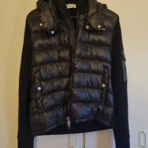 Moncler hybrid jacka - Svart pufferjacka från Moncler med stickade ärmar och quiltad front. Jackan har huva, dragkedja framtill och Moncler-logga på ärmen. Materialmix av polyester och stickad ull ger en snygg och sportig look. Perfekt för kyliga dagar. Jackan är knappt använd och är i storlek S