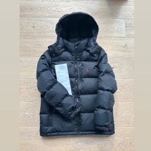 Ralph Laurent jacka - Säljer en svart pufferjacka från Polo Ralph Lauren med huva och dragkedja. Jackan har en klassisk quiltad look och en liten röd logga på bröstet. Perfekt för kalla dagar och riktigt snygg till streetwear-stilen. Perfekt timing att köpa din jacka nu till vintern. Kvitto följer givetvis med och är helt äkta och sprillans ny. Köpt på kidsbrandstore. Nypris 3200, mitt pris 1899. 