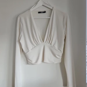 Vit croppad v-ringad topp Bik Bok - Säljer en stilren vit croppad topp från Bik Bok med djup v-ringning och långa ärmar. Toppen har en snygg omlott-effekt framtill och är tillverkad i mjukt material som sitter bekvämt. Perfekt för dig som gillar minimalistisk och trendig stil.