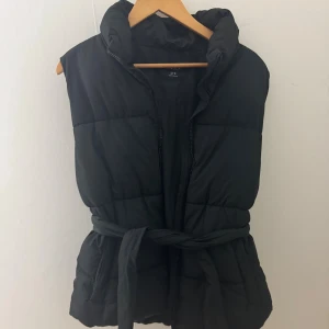 Svart dunväst från Lindex - Svart dunväst från Lindex med quiltad design och hög krage. Västen har dragkedja framtill och ett bälte i midjan för snygg passform. Perfekt att slänga över en hoodie eller stickad tröja när det är kyligt ute. Säljer den då den är stor på mig 