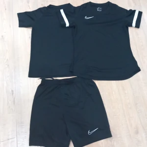 Svarta Nike Dri-Fit träningsshorts och två stycken T-shirts  - Svarta träningsshorts från Nike med klassisk vit swoosh-logga på benet. Tillverkade i lätt och snabbtorkande Dri-Fit polyester, perfekt för sport och träning. Elastisk midja för skön passform och enkel rörelsefrihet.