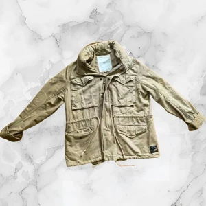 Beige parkajacka från Ralph Lauren - Parkajacka från Denim & Supply Ralph Lauren i beige bomull. Jackan har flera stora fickor framtill, snörning i midjan och patch med amerikansk flagga på ärmen. Klassisk militärinspirerad look med hög krage och detaljerade sömmar.