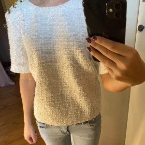 Vit bouclé blus med guldknappar H&M - Säljer en vit bouclé blus från H&M med korta ärmar och tre snygga guldknappar på ena axeln. Blusen har en boxig passform och är tillverkad i ett strukturerat tyg som ger en lyxig känsla. Perfekt att styla med jeans eller kjol för en chic look.