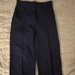 Mörkblå kostymbyxor med pressveck - Snygga mörkblå kostymbyxor med raka ben och tydliga pressveck framtill. Byxorna har hög midja och stängs med knapp och dragkedja. Perfekta för en clean och stilren look. Materialet känns mjukt och har en lätt struktur.