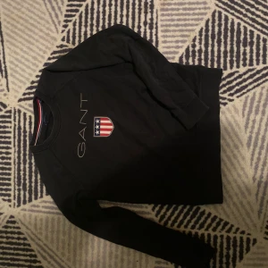 Svart sweatshirt från GANT med logga - Svart sweatshirt från GANT med broderad logga och sköld med stjärnor och ränder på bröstet. Klassisk rund halsringning och långa ärmar. Perfekt för dig som gillar stilren och sportig look.