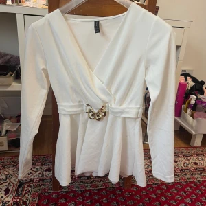 Vit omlottopp med gulddetalj från amisu  - Snygg vit omlottopp från amisu med lång ärm och markerad midja. Toppen har ett brett bälte med chunky guldfärgad kedjedetalj framtill. Mjuk och stretchig känsla, perfekt till jeans eller kjol. V-ringad och lätt utsvängd nertill för en trendig siluett. Toppen har vissa sminkfläckar som visas på bilderna men kanske finns några fler små som jag inte märkt av.