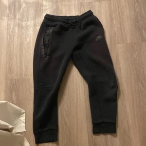 Svarta mjukisbyxor från Nike M - Svarta mjukisbyxor från Nike med resår i midjan och muddar vid bensluten. Byxorna har dragkedjeförsedd ficka på ena sidan och diskret Nike-logga på låret. Perfekta för chill eller träning, tillverkade i mjukt material.