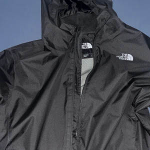 Svart vindjacka från The North Face - Svart vindjacka från The North Face med huva och dragkedja framtill. Jackan har en stilren look med logga på bröstet och är tillverkad i lätt och slitstark polyester. Perfekt för blåsiga dagar och enkel att matcha med allt.