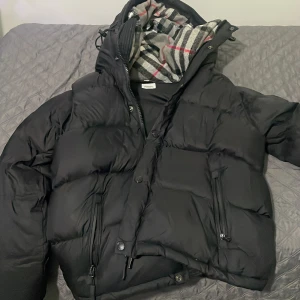 Svart pufferjacka från Burberry - Säljer en svart pufferjacka från Burberry med klassiskt rutigt foder i huvan. Jackan har dragkedja, knappar och två snedställda fickor framtill. Stor Burberry-patch på ärmen och en riktigt fet, varm känsla. Perfekt för kalla dagar när du vill se stilren ut.