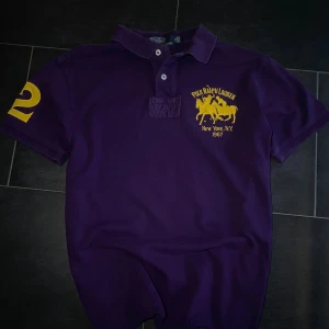 Lila pikétröja Polo Ralph Lauren - Snygg lila pikétröja från Polo Ralph Lauren med gul broderad logga och text på bröstet samt en stor gul siffra 2 på ärmen. Klassisk krage med två knappar och korta ärmar. Perfekt för dig som vill sticka ut med stil.