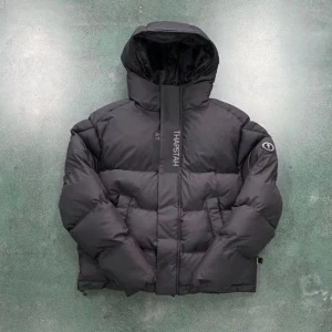 Svart pufferjacka från Trapstar - Svart pufferjacka från Trapstar med stor logga broderad på ryggen och mindre logga framtill. Jackan har huva, dragkedja och två stora fickor. Puffig och varm modell med justerbar nederkant och emblem på ärmen. Perfekt för kalla dagar och streetwear-stil.