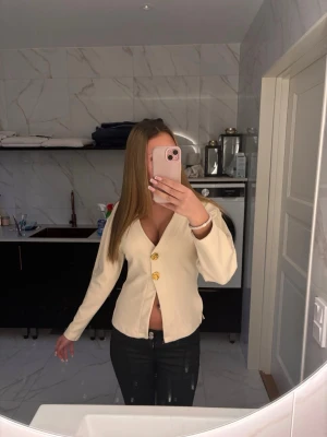 Krämvit kofta med guldknappar - Säljer en krämvit kofta med djup v-ringning och stora dekorativa guldknappar framtill. Koftan har lång ärm och en rak, något kortare passform. Perfekt att styla med jeans eller kjol för en trendig look.