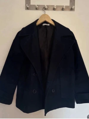 Svart skepparkavaj från Design by S - Klassisk svart skepparkavaj från Design by S med breda slag, dubbelknäppning och rak passform. Jackan har långa ärmar och är tillverkad i ett tjockare material som passar perfekt för kyligare dagar.