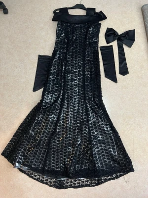 Svart glittrig långklänning med rosett - Elegant svart långklänning med glittrigt mönster, bred krage och tillhörande satinhandskar och stor rosett. Klänningen har en figurnära passform och är perfekt för bal eller fest. Materialet är spets och polyester med paljettdetaljer för extra glamour.