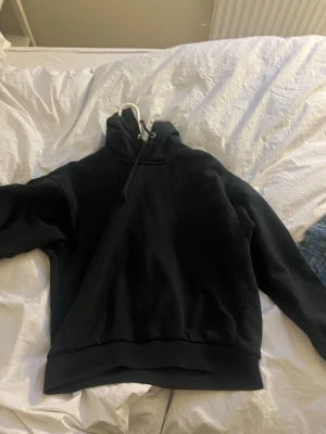 Svart hoodie från 157 med huva - Säljer en svart hoodie från 157 med klassisk huva och dragsko. Hoodien har en enkel och clean design, ribbade muddar och är gjord i mjukt sweatshirtmaterial. Perfekt för dig som gillar en avslappnad och stilren look.